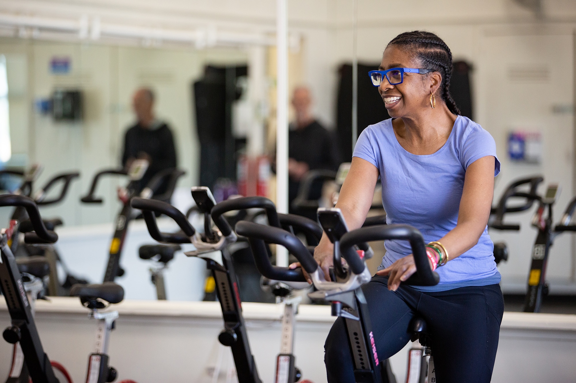 A woman wearing glasses and a purple shirt smiles while using a stationary exercise bike in a gym with mirrors and other bikes visible in the background.