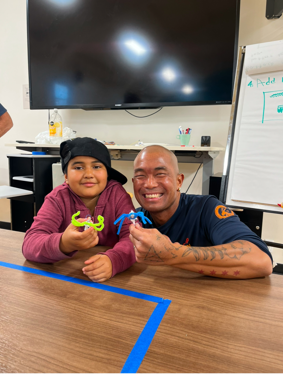 A smiling adult and child sit at a table holding colorful plastic models, celebrating National Mentoring Month. A TV, whiteboard, and markers can be seen in the background, highlighting the impact of programs like Reach & Rise.