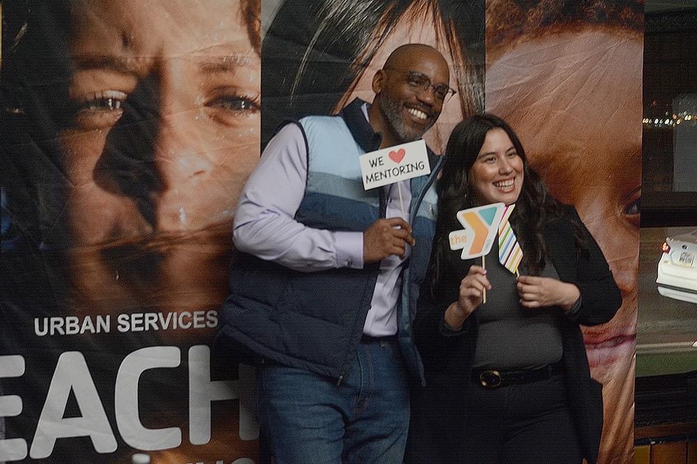 Two people stand together indoors, smiling and holding signs that read "We ♥ Mentoring" and "the Y," in front of a banner with faces and the words "Urban Services," celebrating Reach & Rise during National Mentoring Month.