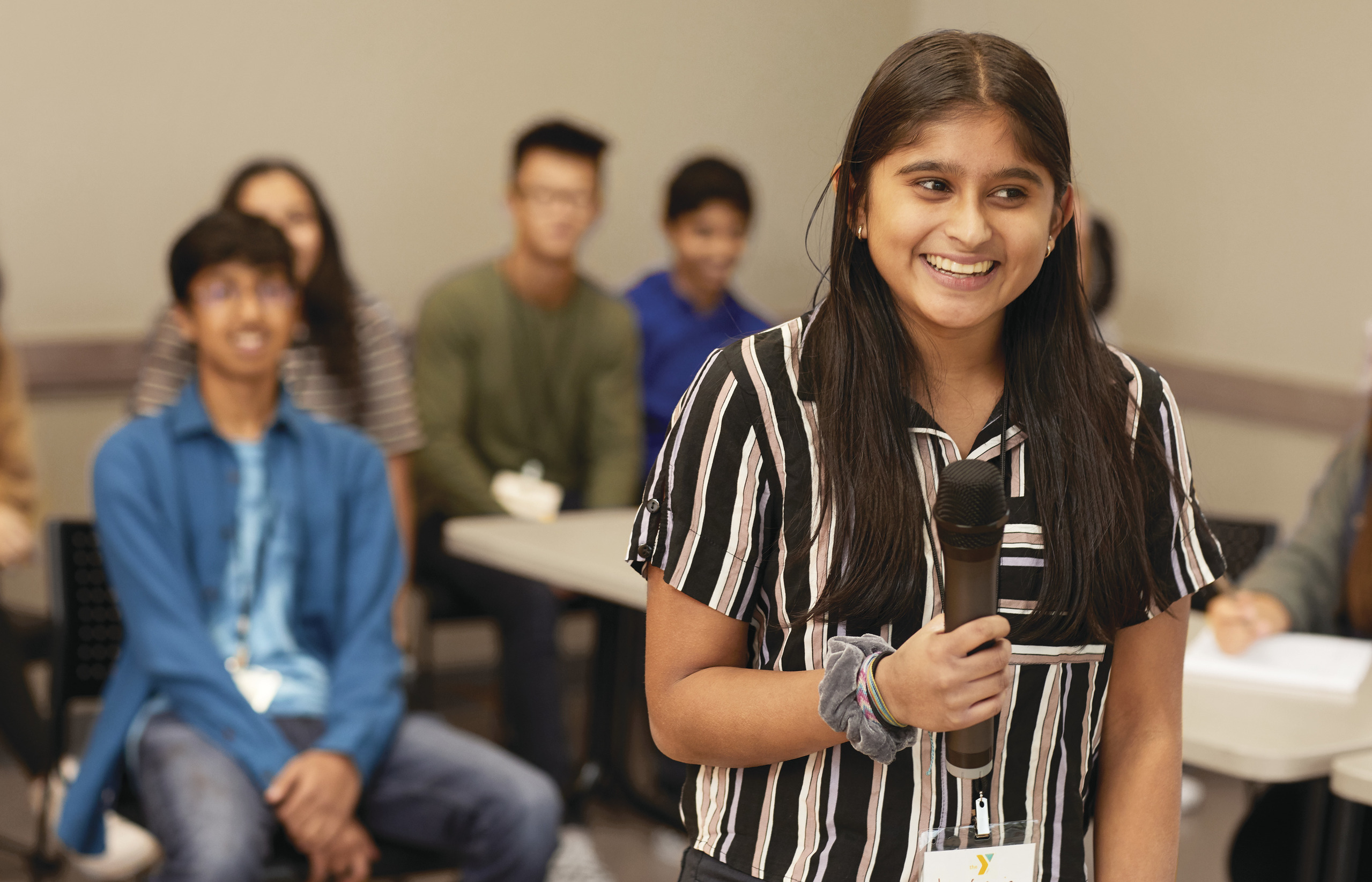 Teen girl with long hair holding a microphone and other teens in the background.