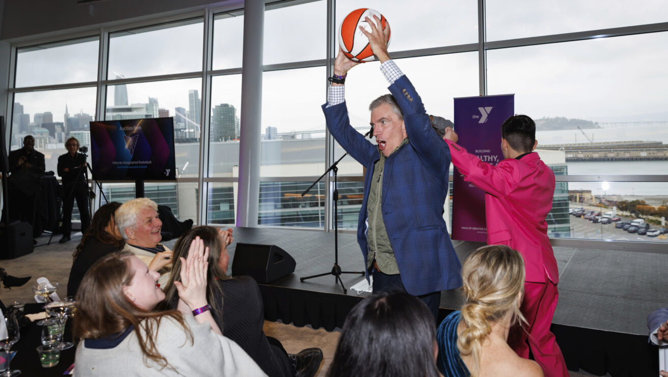 A man in a blue suit holds a basketball over his head in front of an applauding audience at the Y Gala, an indoor event with large windows and stunning city views.