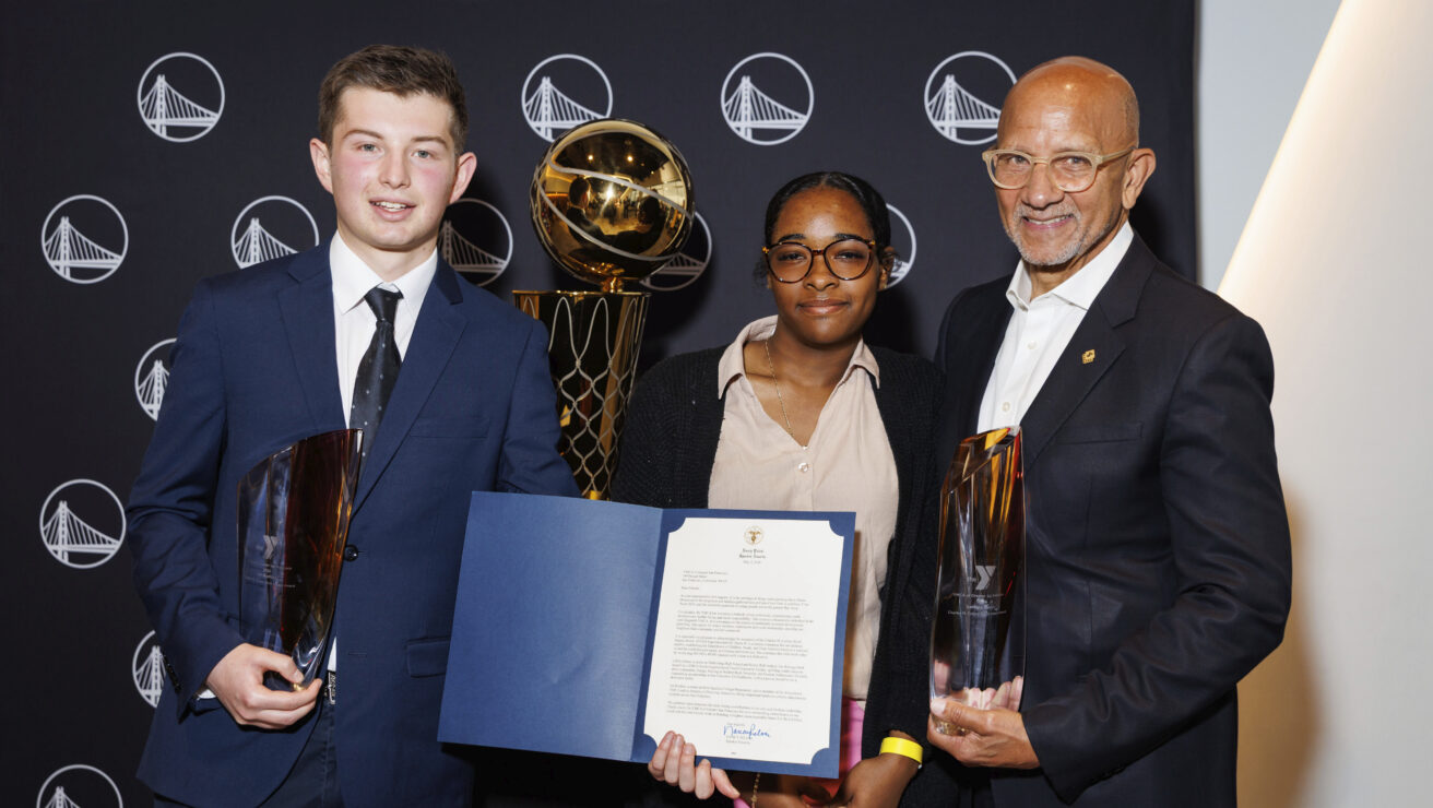 Three people in formal attire pose with trophies and a certificate in front of a backdrop featuring a repeated basketball logo and a large gold trophy, capturing the celebratory spirit of the Y Gala.
