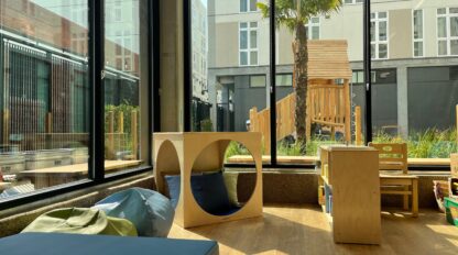 Sunny daycare room with large windows, soft seating, wooden play furniture, and a view of an outdoor playground with a palm tree—experience this inviting space during our preschool open house at the Tung Lok Early Childhood Learning Center.