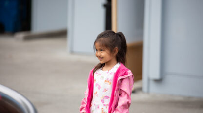 A young girl in a pink jacket and white dress with colorful patterns stands outside, smiling, with a gray building in the background—capturing the joyful spirit of the upcoming preschool open house at Alice Chalmers Preschool.