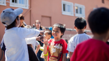 A group of children stand and interact outdoors near a building during the Open House: Mission YMCA Preschool; one boy in a red shirt smiles and waves at the camera.