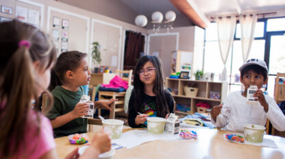 Four children sit at a table in a classroom, eating snacks and drinking from milk cartons during the Open House: Mission YMCA Preschool.