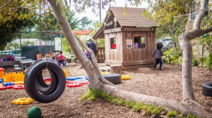 Outdoor playground with a tire swing, large foam building blocks, and a wooden playhouse. Children are playing near the playhouse under trees during our preschool open house, where families can explore and experience the joyful environment firsthand.