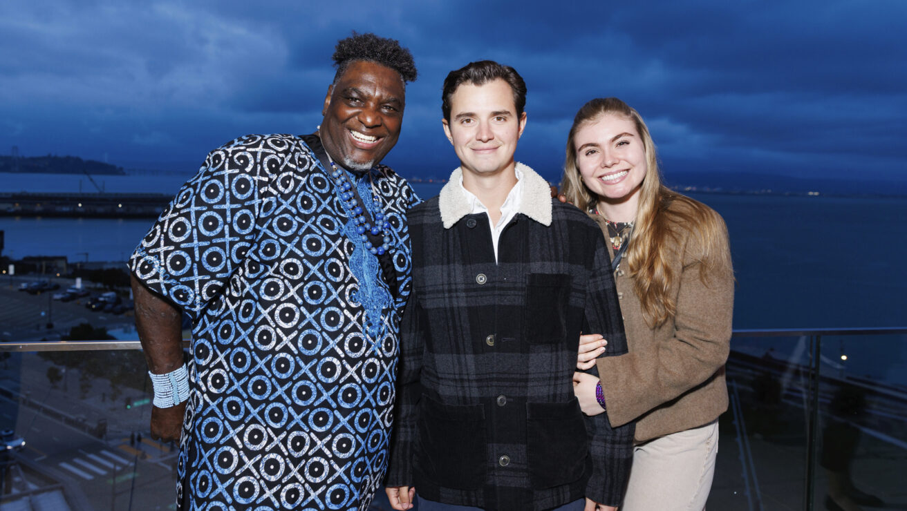 Three people stand together and smile for a photo on a balcony overlooking water, with a cloudy evening sky in the background.