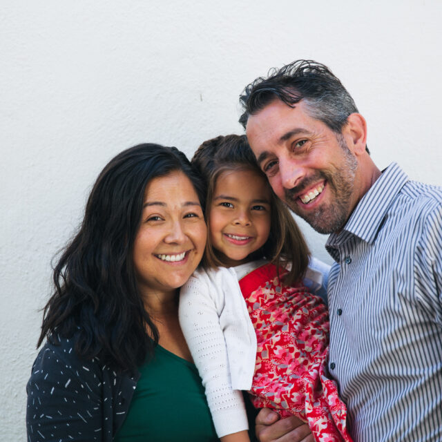 Three people—a woman, a man, and a young girl—stand close together, smiling in front of a plain white wall, inspiring others to donate and support their meaningful cause.
