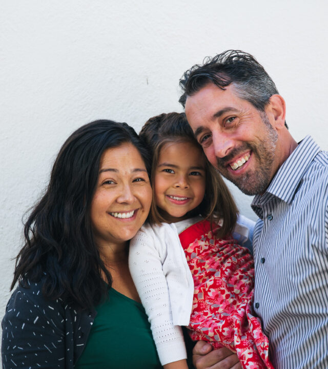 Three people—a woman, a man, and a young girl—stand close together, smiling in front of a plain white wall, inspiring others to donate and support their meaningful cause.