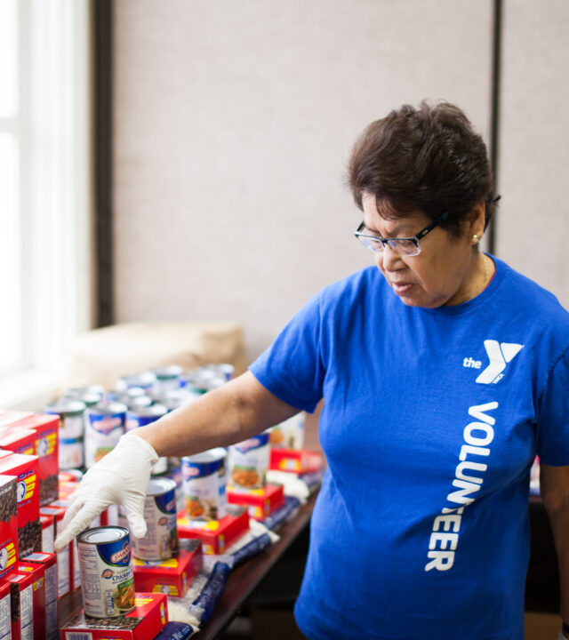A person wearing a blue “volunteer” shirt and gloves organizes canned goods and boxed food items on a table indoors, encouraging others to donate and support those in need.