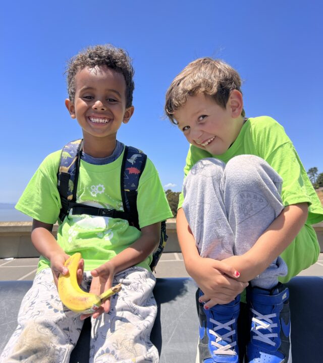 Two boys in bright green YMCA San Francisco t-shirts smiling at the camera, one holding a banana.