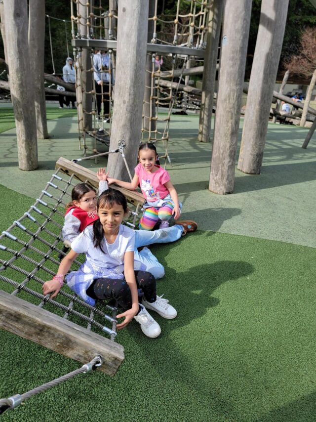 Three children from the Mission YMCA school year camp sit on a rope climbing structure at a playground.