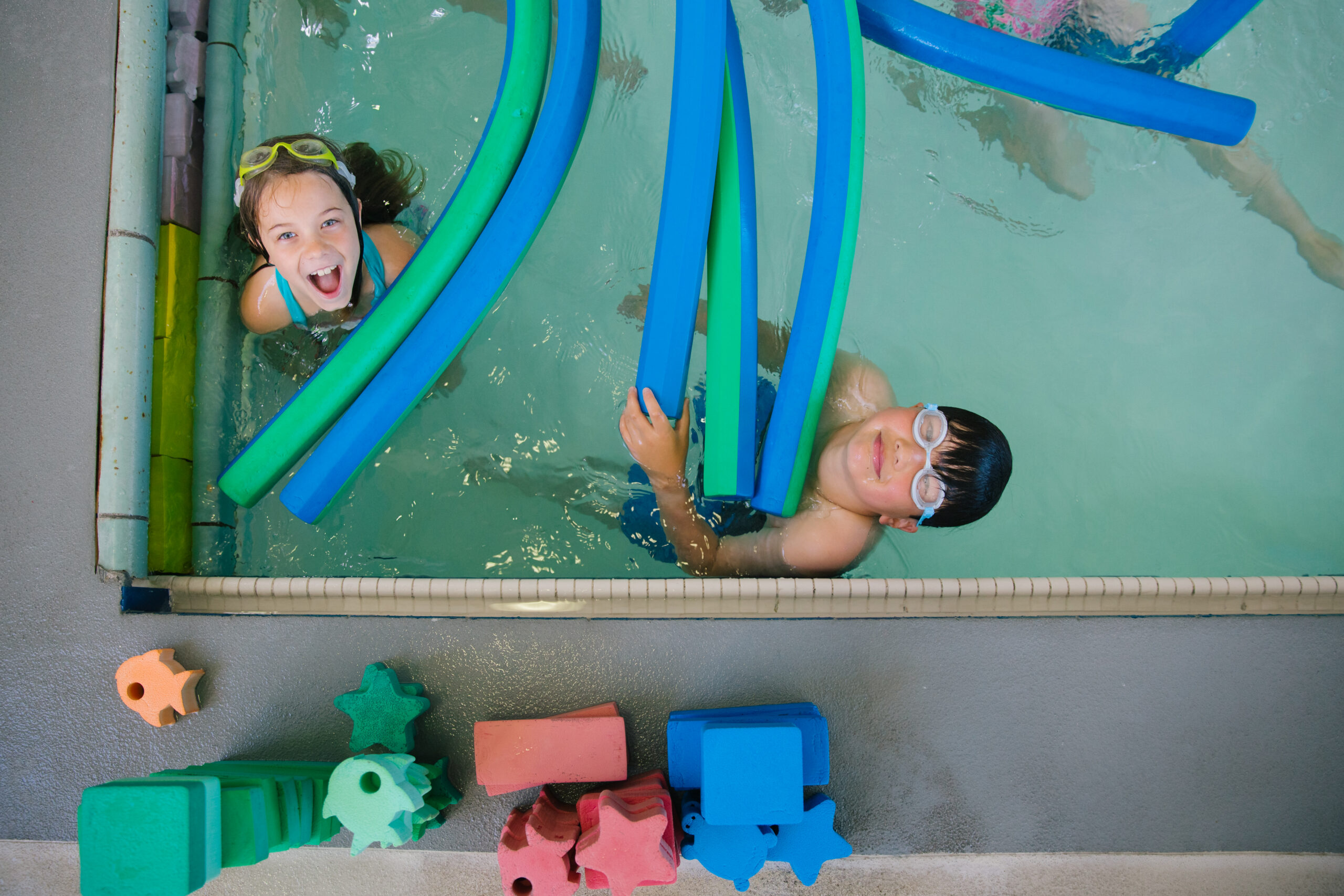 Two children wearing swim goggles play with blue pool noodles in a Presidio Community YMCA pool, with foam toys sitting on the deck.