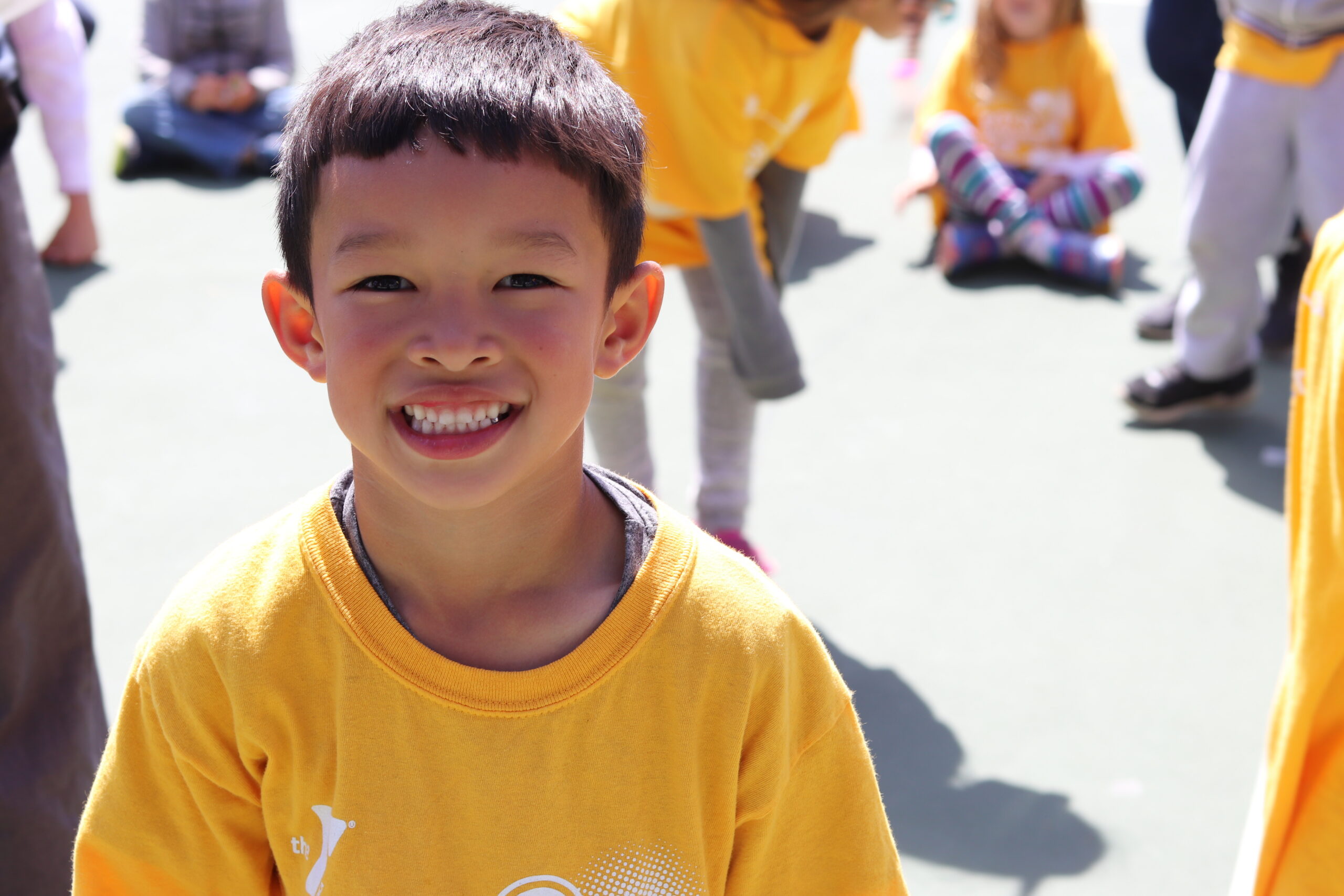 A young boy in a yellow t-shirt smiles at the camera during Chinatown YMCA School Year Camp, with other children sitting and standing in the background.