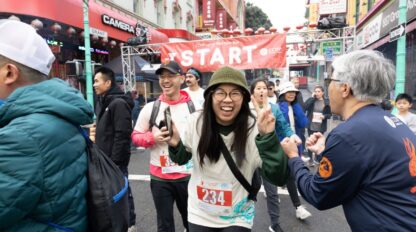Runners at the start line of the CCHP Lunar New Year Run, reflecting the community history of San Francisco as a woman in the center smiles and makes a peace sign.