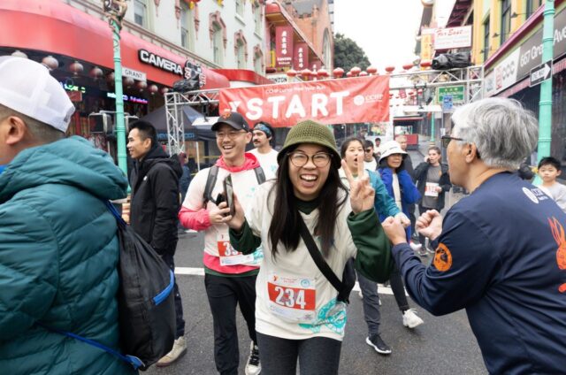 Runners at the start line of the CCHP Lunar New Year Run, reflecting the community history of San Francisco as a woman in the center smiles and makes a peace sign.