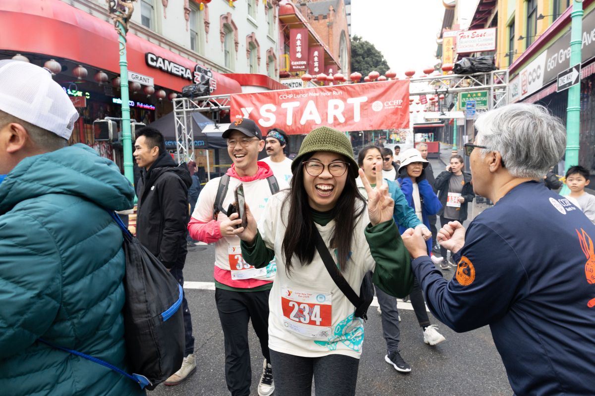 Runners at the start line of the CCHP Lunar New Year Run, reflecting the community history of San Francisco as a woman in the center smiles and makes a peace sign.