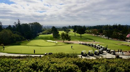 A Bayview STEAM golf course with a putting green in the foreground, parked golf carts, and a line of trees extending toward the horizon under a cloudy sky.