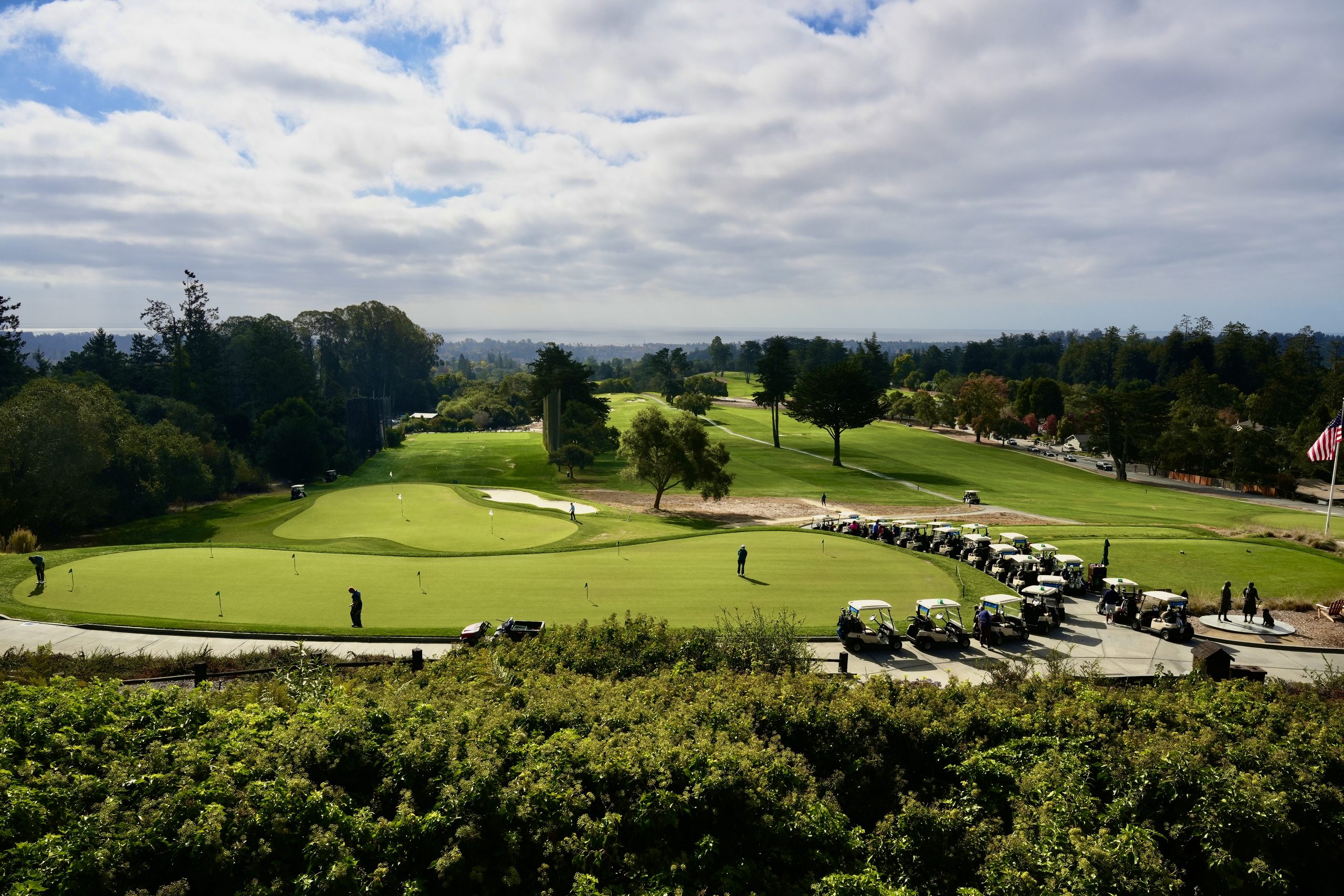 A Bayview STEAM golf course with a putting green in the foreground, parked golf carts, and a line of trees extending toward the horizon under a cloudy sky.